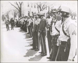 Black and white photo of a line of police officers wearsing gas masks. Two of the police officers look at the camera.